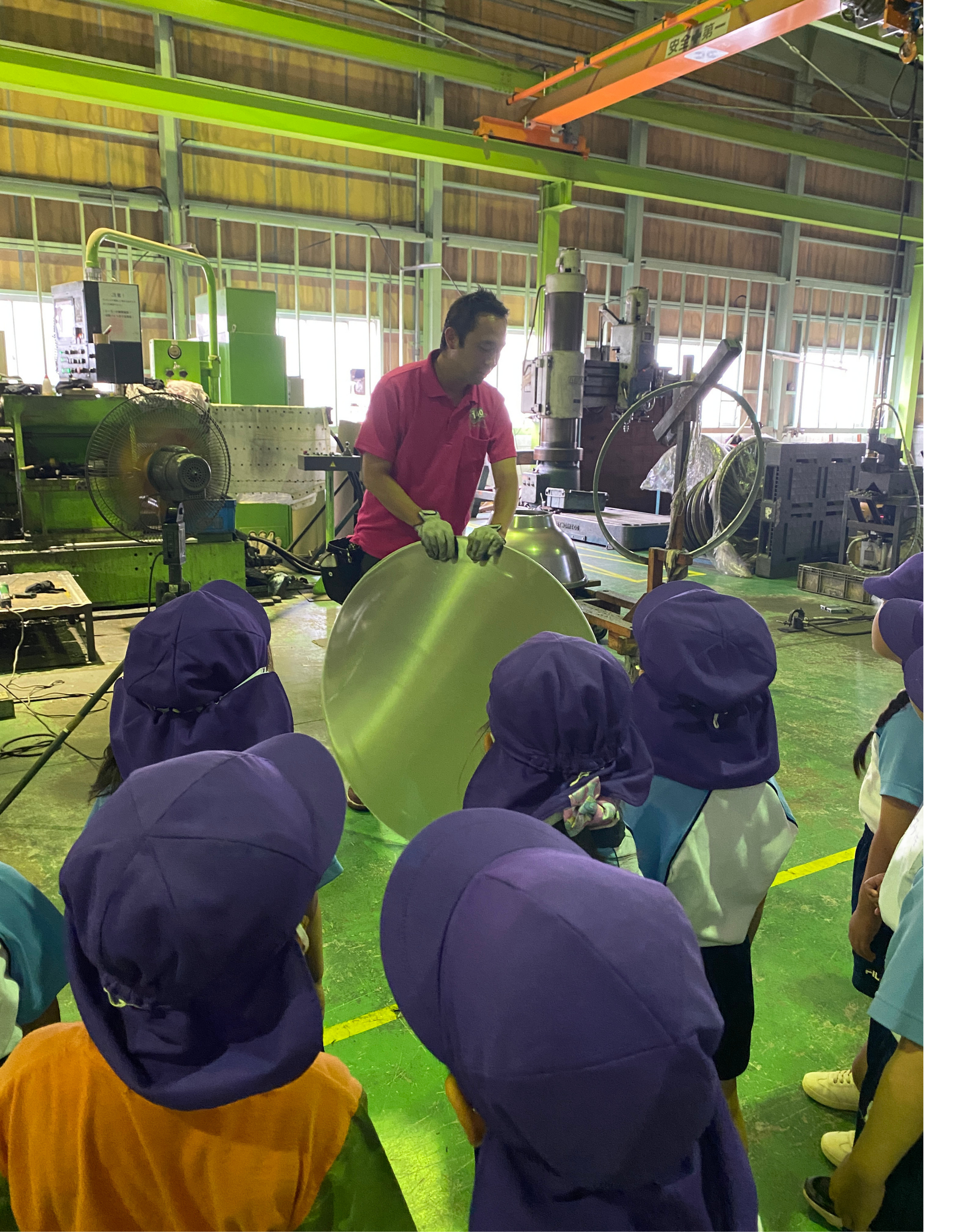 服部工業小豆坂工場にある国内でも希少なスピニングマシンの前で、めぐみ幼稚園の園児たちに回転釜のお釜部分がどんな材料からどのように作られるかを分かりやすく説明しているスタッフの様子